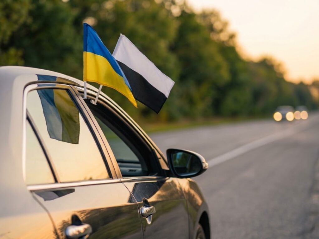 Чорна машина на дорозі між лісосмугами, на ній державний поапор і прапор надії