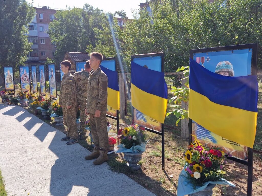 Хлопець у військовій формі знімає прапори з банерів, поруч - почесна варта зі ще двох хлопців у військовій формі