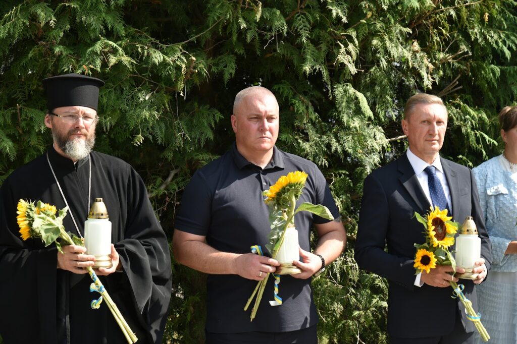 три чоловіка на відкритті алеї пам'яті тримають в долонях квіти та лампадки