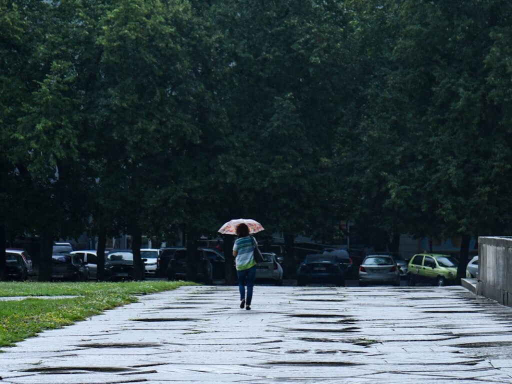 Жінка під парасолькою йде по доріжці з бетонних плит до стоянки машин під деревами