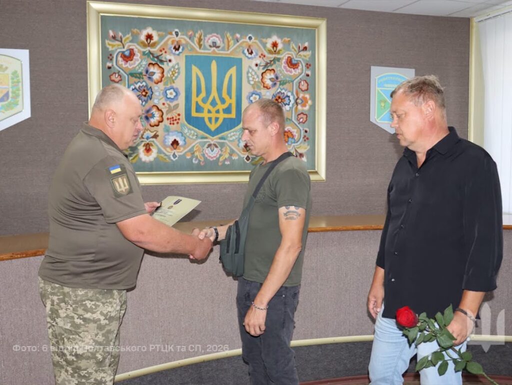Військовослужбовець ТЦК вручає колишньому воїну нагороду, поряд чекає чоловік, щоб вручити нагородженому троянду