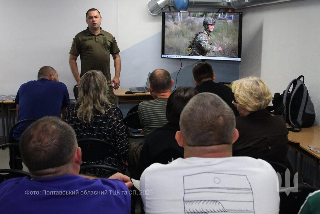 Укриття, військовик виступає перед людьми в залі, поряд із ним екран, на якому людина у військовій формі долає смугу перешкод