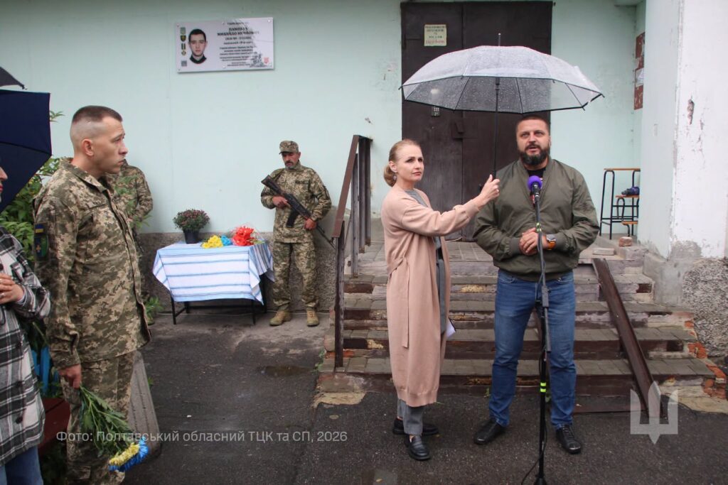військовослужбовець перед мікрофоном, ведуча тримає над ним парасольку, на задньому плані на стіні будинку меморіальна дошка, під нею стіл із квітами, поруч вхід у під'їзд