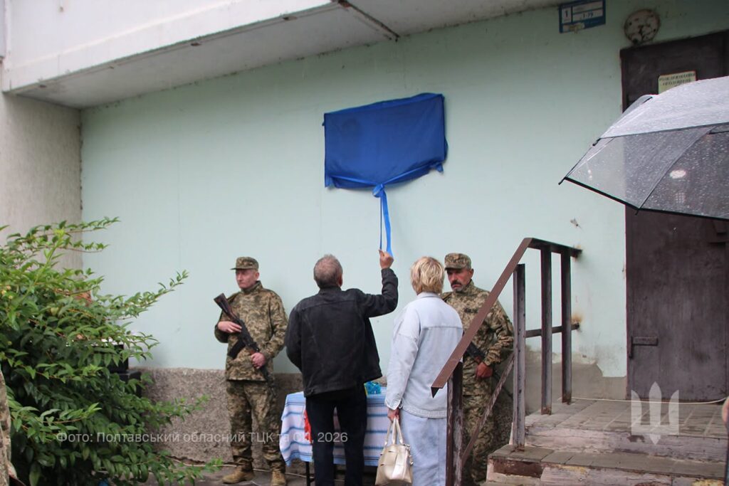 чоловік та жінка знімають синю тканину з меморіальної дошки на стіні будинку, поряд у почесній варті стоять двоє військовиків