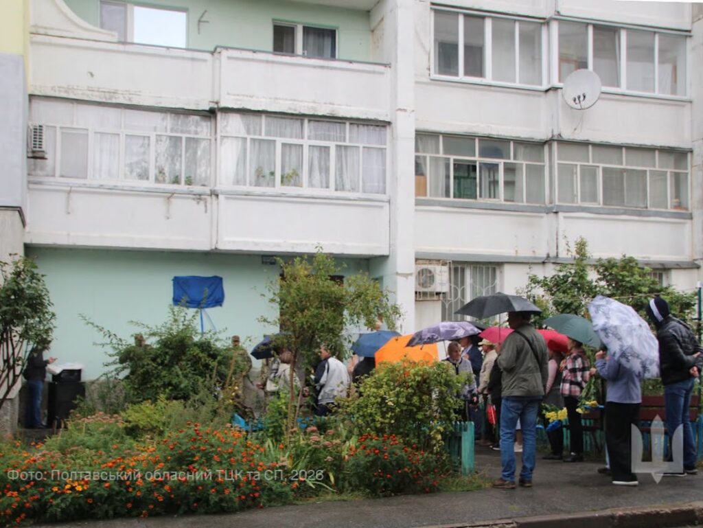 люди під парасольками на подвір'ї біля багатоповерхівки, на якій закрита синьою тканиною меморіальна дошка