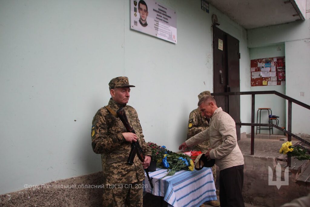 чоловік кладе квіти до меморіальної дошки, у почесній варті поряд стоять двоє військовиків з автоматами