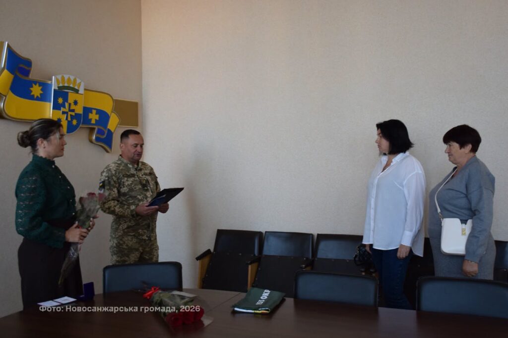у залі засідань військовик зачитує документ родині загиблого воїна, поряд стоїть жінка з квітами