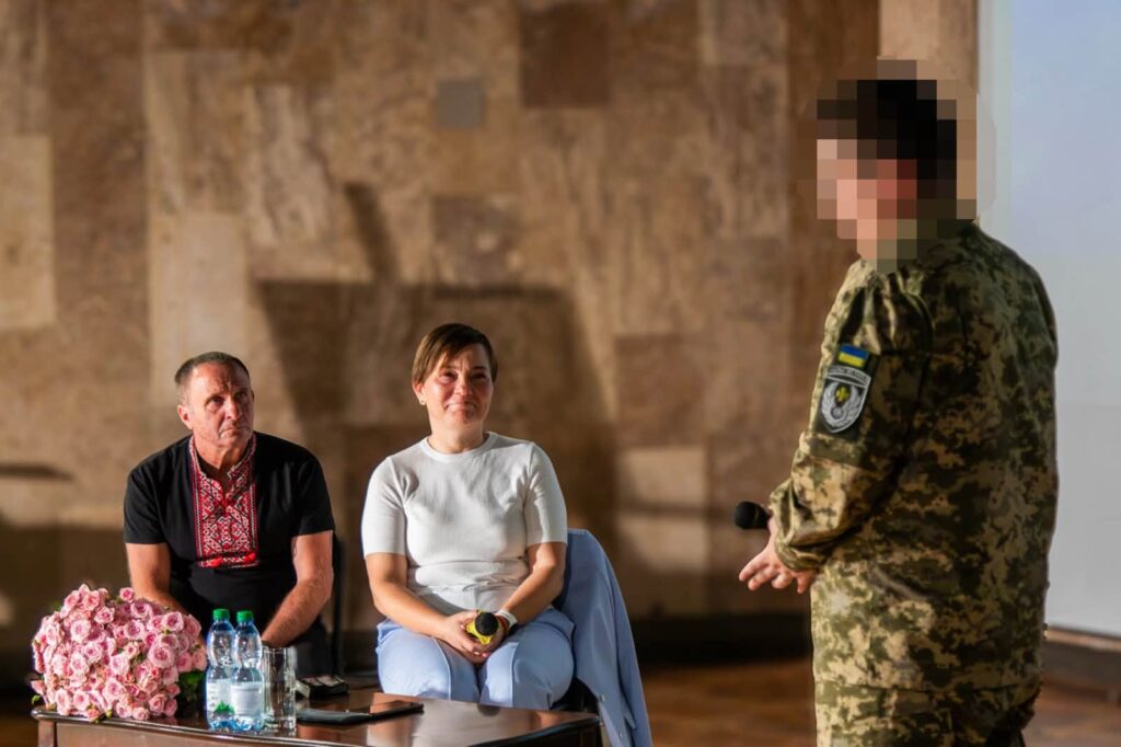 Військовик стоїть біля чоловіка та жінки, які сидять на стільцях біля екрану