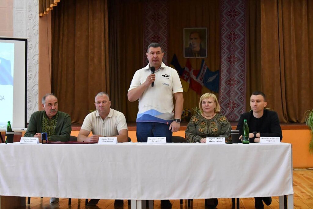стіл президії, за ним троє чоловіків і жінка сидять, один стоїть і говорить у мікрофон