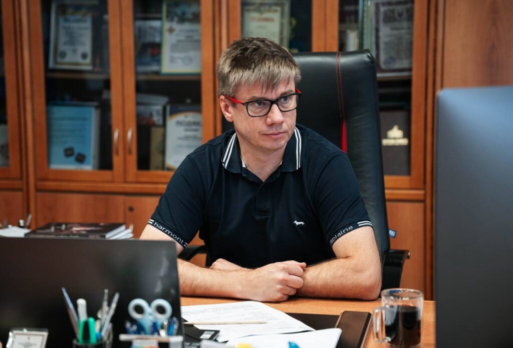 Чоловік у темній футболці сидить за столом перед монітором, поруч ноутбук і канцелярське приладдя, позаду - шафа з документами та іншими предметами