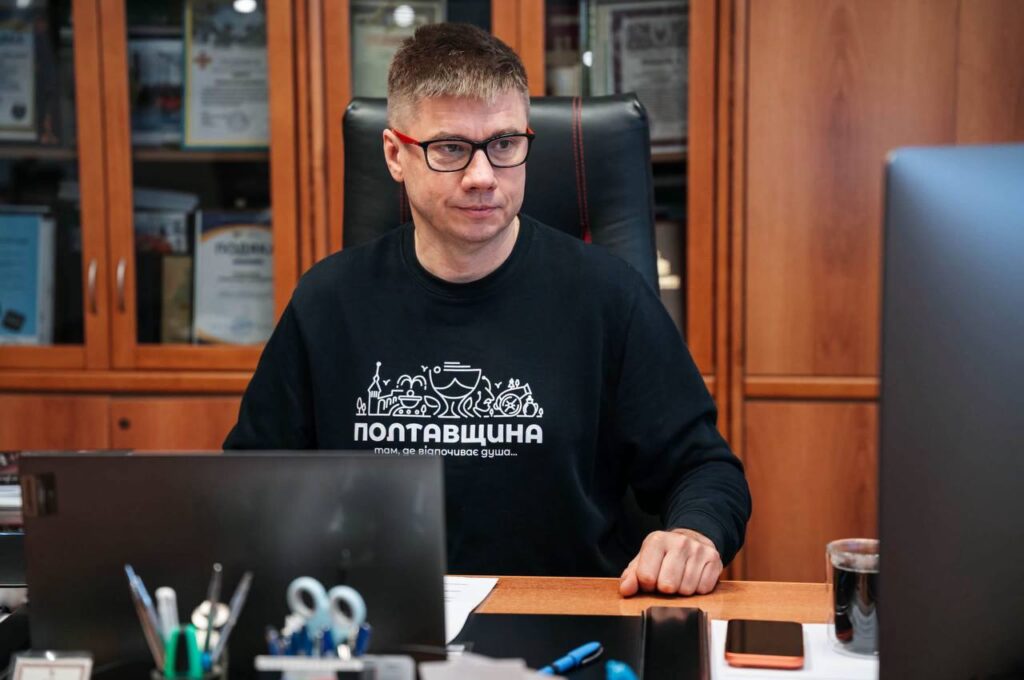 Олександр Біленький у чорному світшоті з написом Полтавщина сидить за столом, на якому стоїть ноутбук, екран, лежить канцелярське приладдя, позаду нього - шафа з документами та нагородами