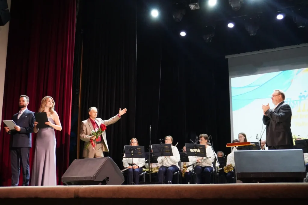 На сцені: чоловік і жінка у вечірньому вбранні - ведучі, співак із квітами, оркестр і диригент, який аплодує
