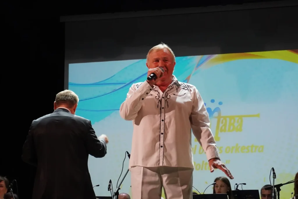 Віктор Щербаков співає на сцені, позаду нього диригент, який керує оркестром, та екран із назвою колективу