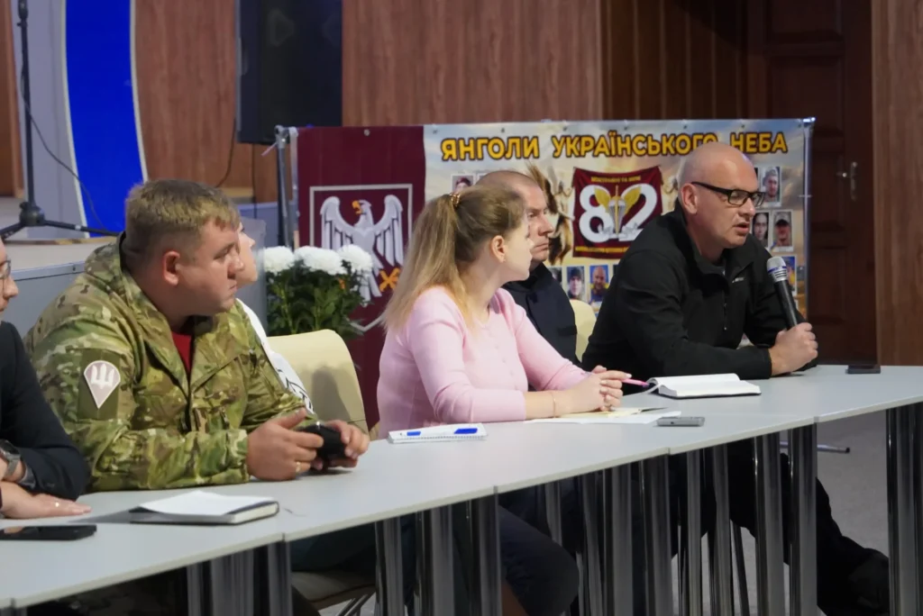 Євген Кончаковський із мікрофоном на тлі банера "Янголи українського неба", поряд із ним за столом сидять люди