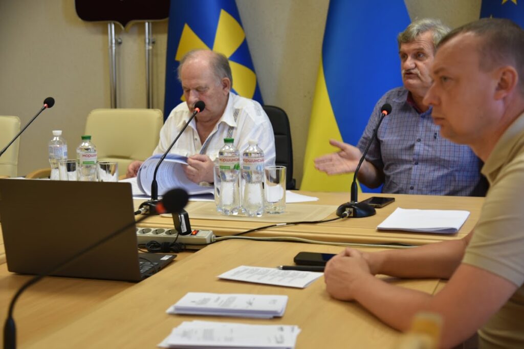 депутати обговорюють питання, на столах перед ними документи, ноутбук, вода, мікрофони
