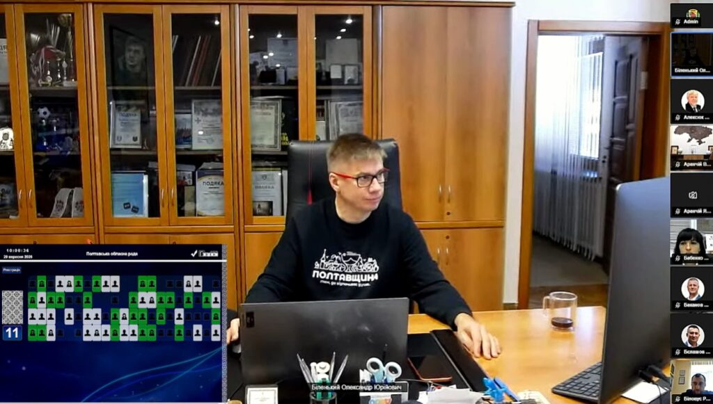 Олександр Біленький у чорному світшоті з написом Полтавщина сидить за столом, на якому стоїть ноутбук, екран, лежить канцелярське приладдя, позаду нього - шафа з документами та нагородами, зліва внизу зображення відображається екран із даними голосування