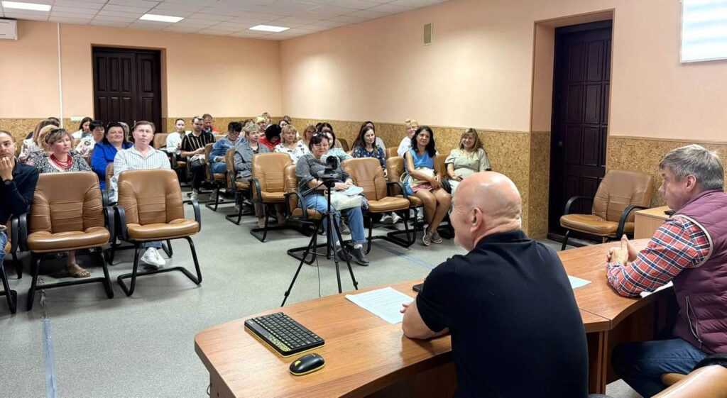 актова зала академії, у ній сидять люди, за столом перед ними Зелюк і Біленький