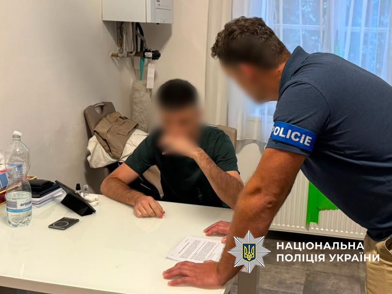 Чоловік у темній футболці сидить за столом і тримає одну руку біля рота, обличчя розмите, над ним "нависає" чоловік у синій футболці-поло з пов'язкою із написом чеською "поліція"
