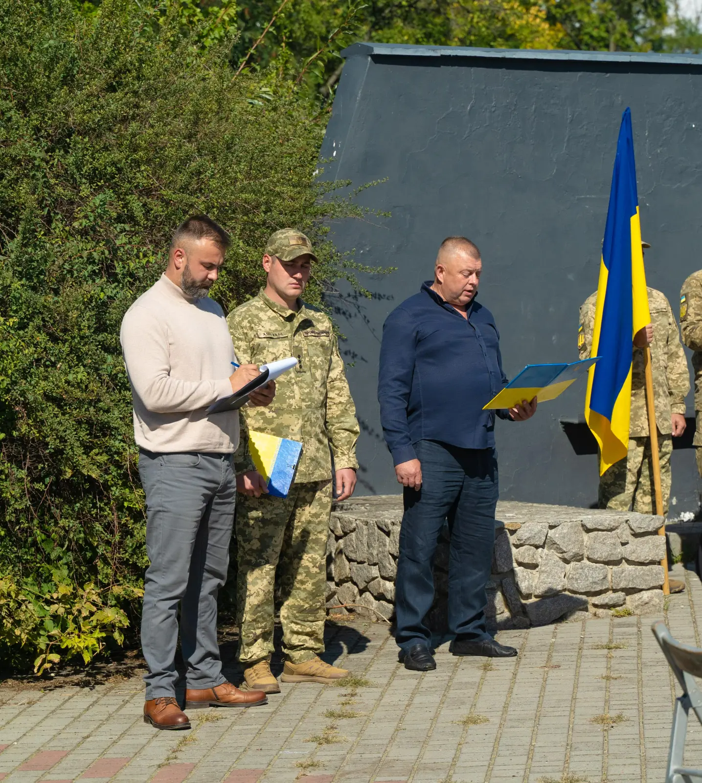 двоє чоловіків у цивільному одязі та один у військовому стоять біля прапора України