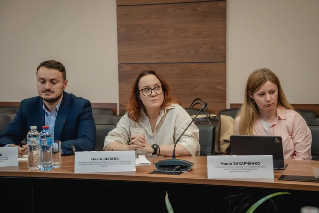 чоловік та дві жінки сидять за столом. перед ними таблички з іменами, прізвищами, посадами