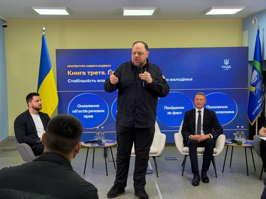 Руслан Стефанчук розповідає про третю книгу Цивільного кодексу, у кріслах позаду нього видять ведучий і Володимир Онищенко, встановлені прапори на екран із презентацією