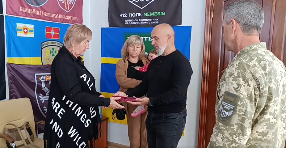 Чоловік у темному цивільному одязі передає нагороду жінці в чорному одязі, поряд стоять жінка з маленькою дитиною на руках і чоловік у військовій формі, все відбувається на тлі різних прапорів