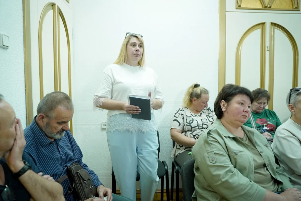 Жінка стоїть біля стіни і говорить, біля неї сидять на стільцях інші люди
