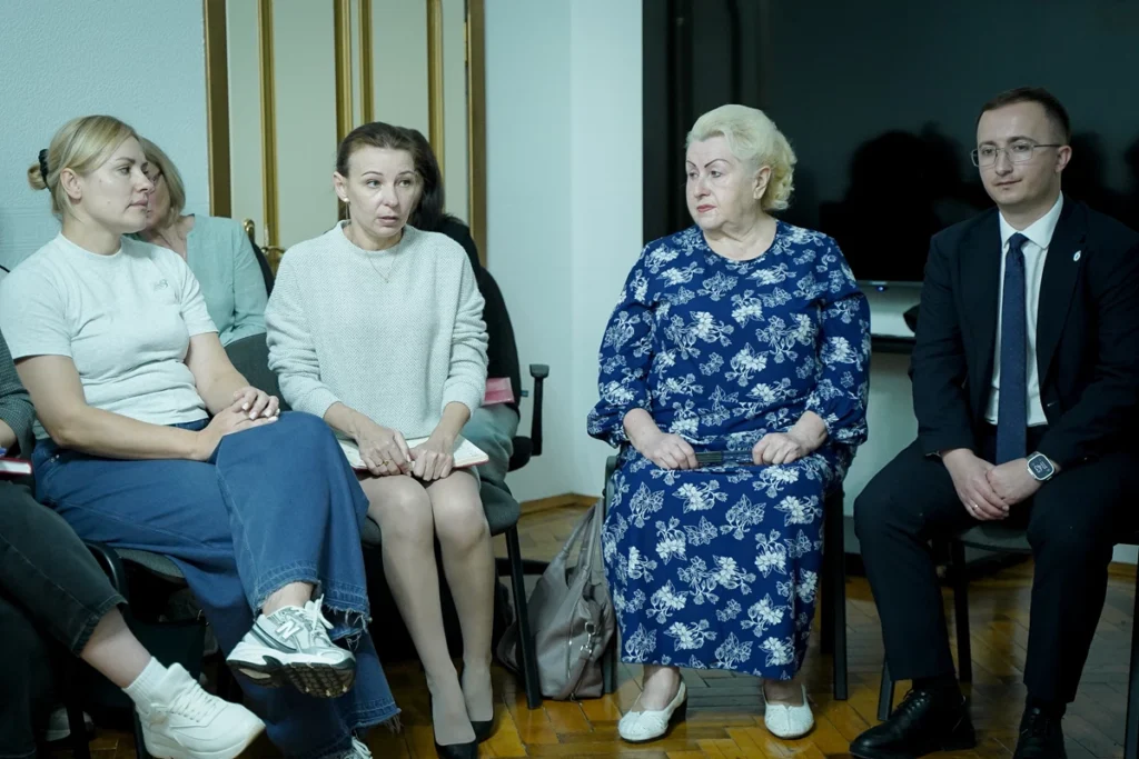 люди сидять на стільцях навпроти екрану, одна з жінок говорить 