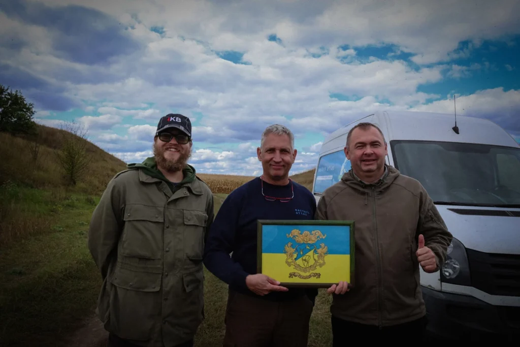 троє чоловіків біля машини в полі, один тримає подарунок із українською символікою
