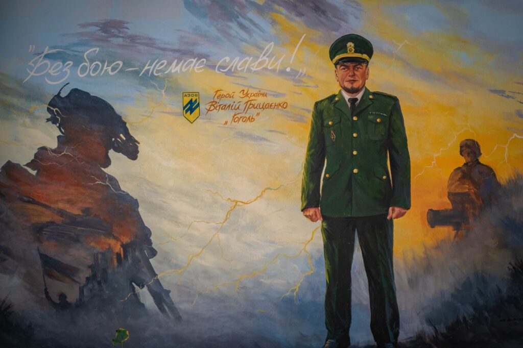 На муралі зображено Героя України Віталія Грицаєнка з позивним «Гоголь». У правій частині стінопису у повний зріст стоїть сам Віталій у зеленій парадній військовій формі з кашкетом та золотими ґудзиками. Ліворуч від нього на тлі тривожного неба з блискавками та вогняно-помаранчевим заходом сонця виведено розмашистий напис: «Без бою – немає слави!». Трохи нижче розміщено емблему підрозділу «Азов» і підписано його ім'я та позивний. З лівого боку картини темним силуетом вимальовуються руїни будівлі, що символізують бої за Маріуполь, на тлі яких майорить український прапор. У правій частині, за спиною воїна, видніється силует побратима біля бойової позиції. Загальна атмосфера зображення поєднує тему запеклих боїв, стійкості та світлої пам'яті про полеглого захисника.