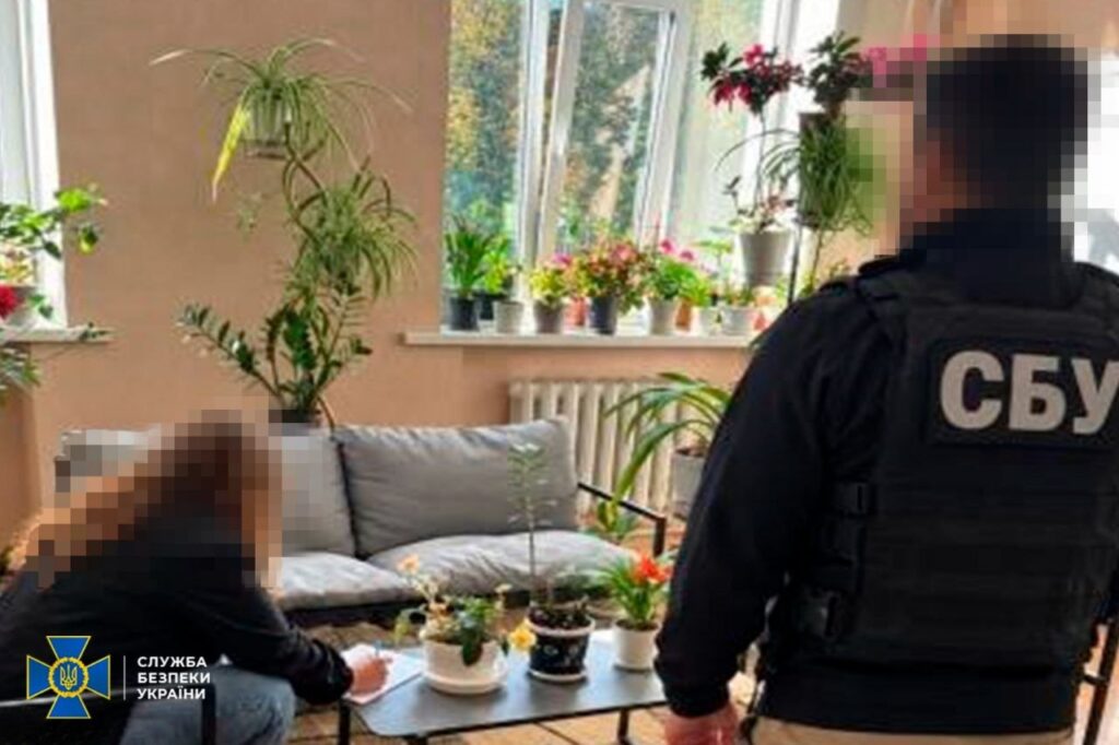 жінка, сидячи за журнальним столиком, біля дивану, у кімнаті з багатьма квітами, оформлює документи, поряд стоїть чоловік в одязі з написом сбу