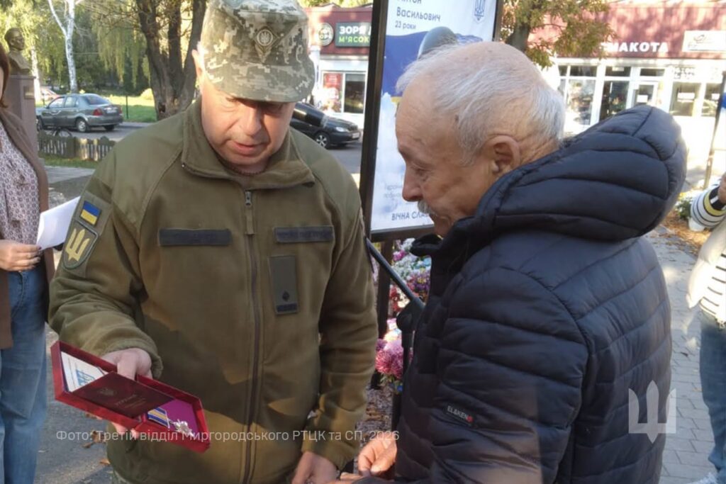 на алеї слави військовослужбовець вручає сивому чоловікові в темній куртці нагороду в червоній коробочці та свідоцтво