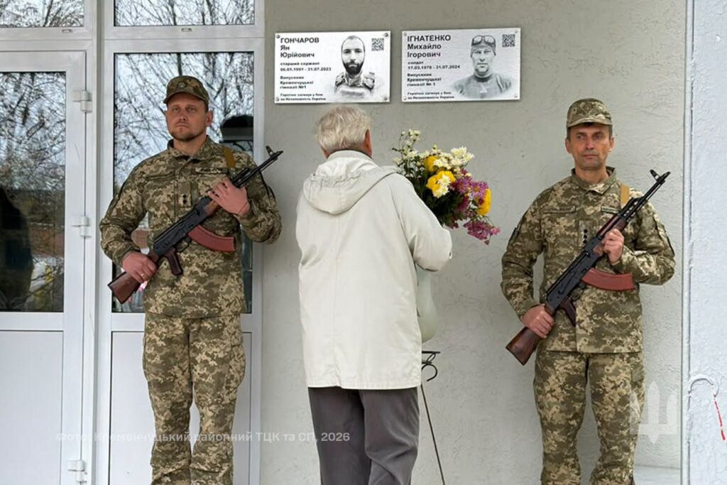 жінка у світлій куртці несе квіти до відкритої меморіальної дошки, біля якої стоїть почесна варта з двох військовиків