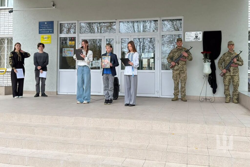 біля входу в будівлю стоять педагоги й школярі, які розповідають про полеглого, біля закритої чорною тканиною меморіальної дошки стоїть почесна варта з двох військовослужбовців