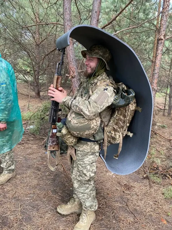 Артем у військовій формі зі зброєю, тримає над собою скатку-каремату, ховаючись від дощу