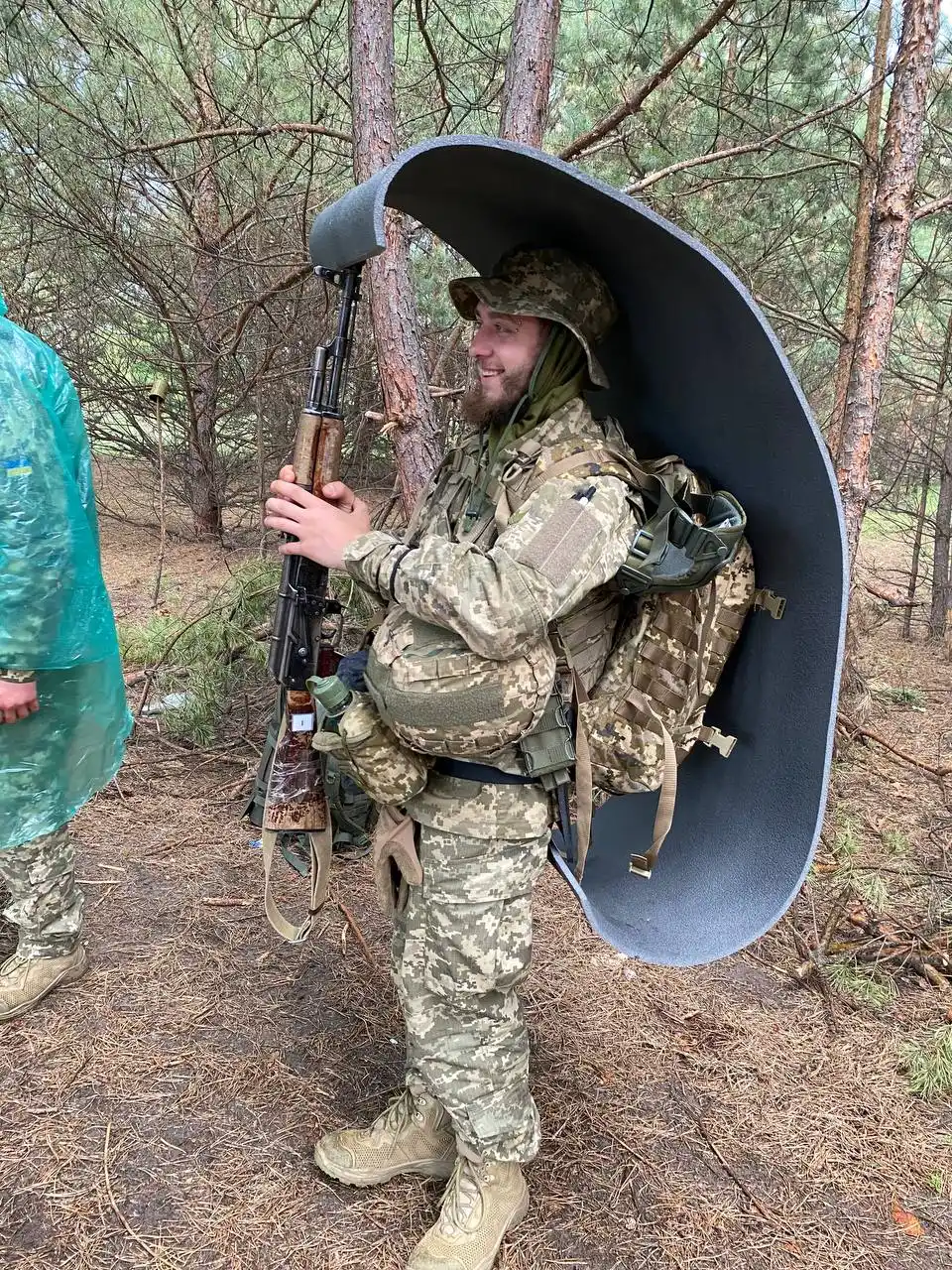 Артем у військовій формі зі зброєю, тримає над собою скатку-каремату, ховаючись від дощу