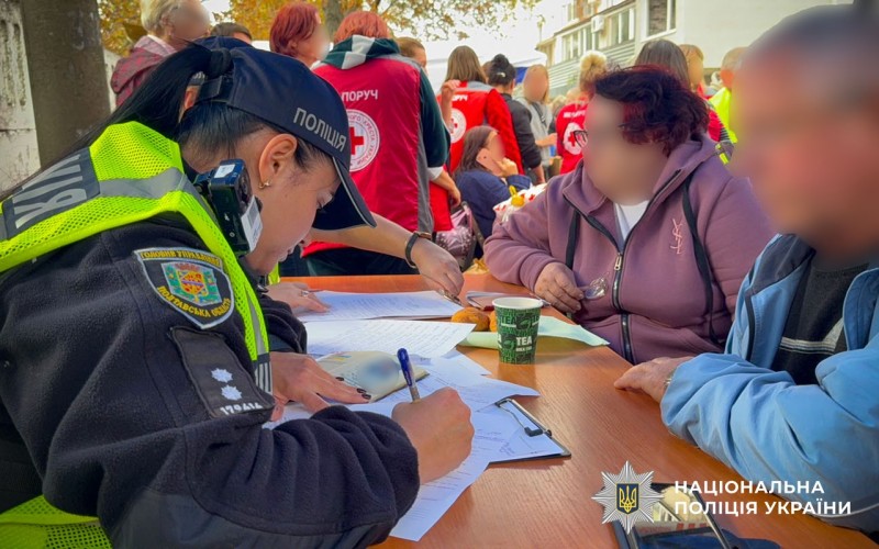 поліцейська за столом оформлює заяву від потерпілих жінки та чоловіка, на задньому фоні стоять люди в спецодязі з червоним хрестом
