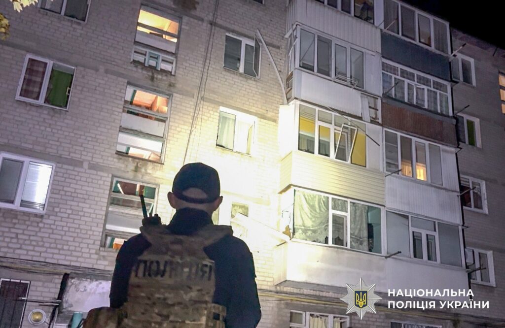 чоловік у формі та бронежилеті з написом поліція дивиться на житловий будинок, пошкоджений обстрілом