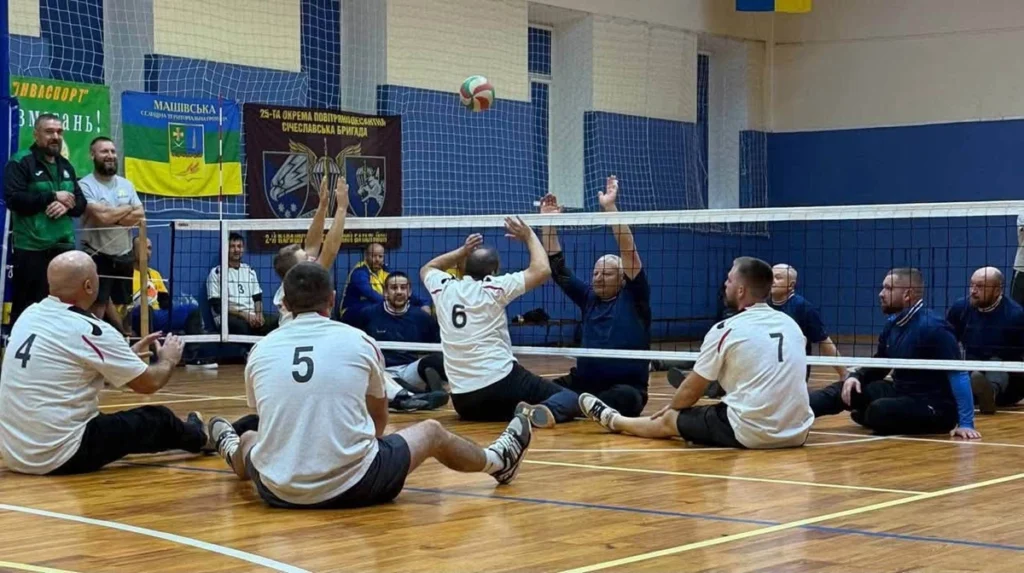 спортсмени в білих та темних футболка сидять на підлозі, м'яч летить над сіткою
