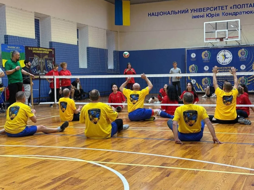 спортсмени в жовтих футболках грають проти спортсменів у червоних футболках, перекидаючи м'яч через сітку сидячи на підлозі